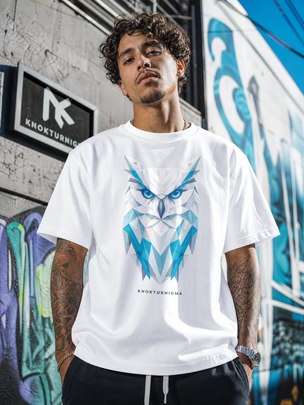 A man wearing a white short-sleeve t-shirt with a blue geometric owl graphic on the front, standing in front of a graffiti-covered wall. The brand name 'Knokturnigma' is printed below the graphic.