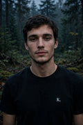A man with short brown hair and light stubble, wearing a black crewneck t-shirt with a small white 'K.' logo on the left chest, standing in a misty, dark forest.